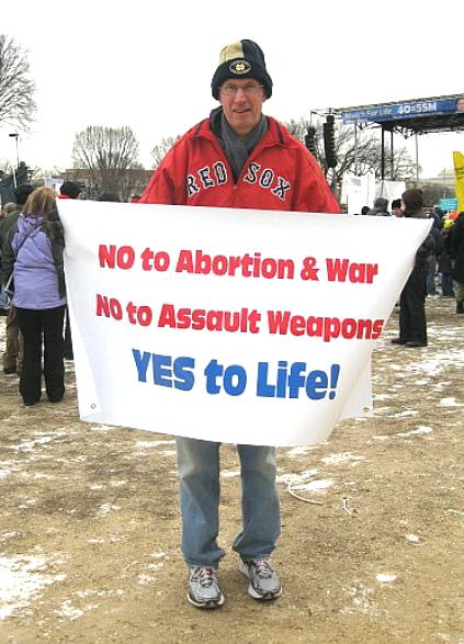 Man holds banner: 'NO to Abortion & War/NO to Assault Weapons/YES to Life!'
