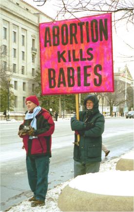 Man, standing on snowy sidewalk, with sign: 'Abortion Kills Babies'